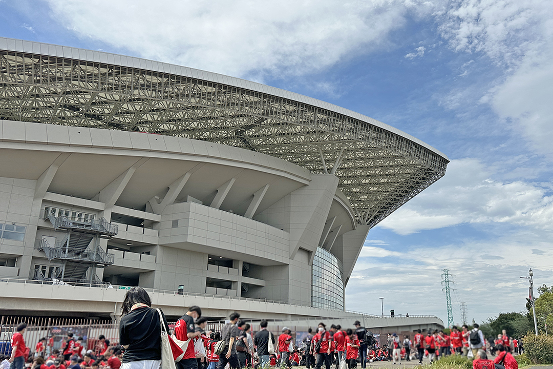 スタジアムの外観写真。空も晴れ、スタジアム周辺には赤いユニフォームを着た多くの人が集まり、仲間と語りあったりしている。試合前のワクワクした雰囲気が伝わってくる。