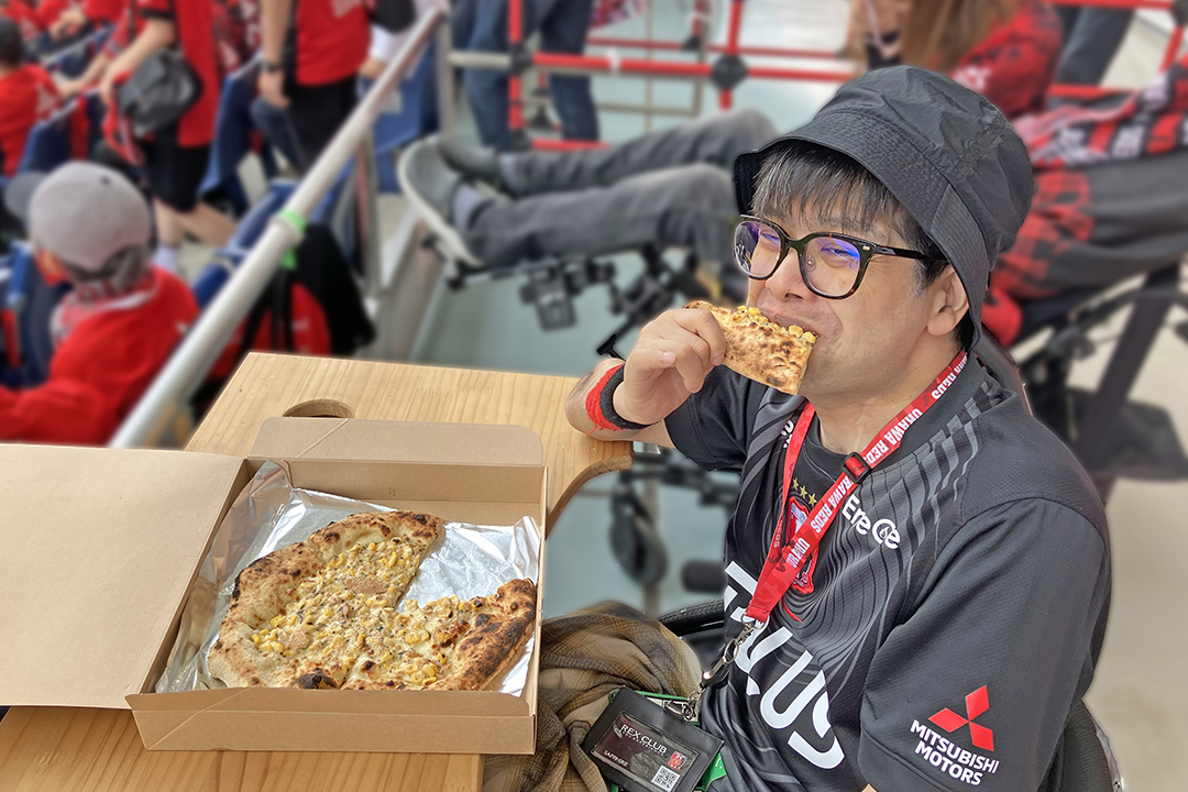 車いす席で、ピザを食べながらサッカー観戦を楽しんでいる翔太。グルメと応援を同時に楽しんでいます。