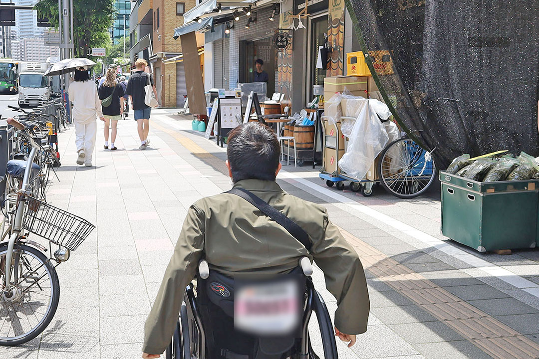 人々がゆったりと歩道を歩いており、道沿いには自転車や店舗、商品が並び、街の活気も感じられ、心地よい街歩きの一場面といった印象です。