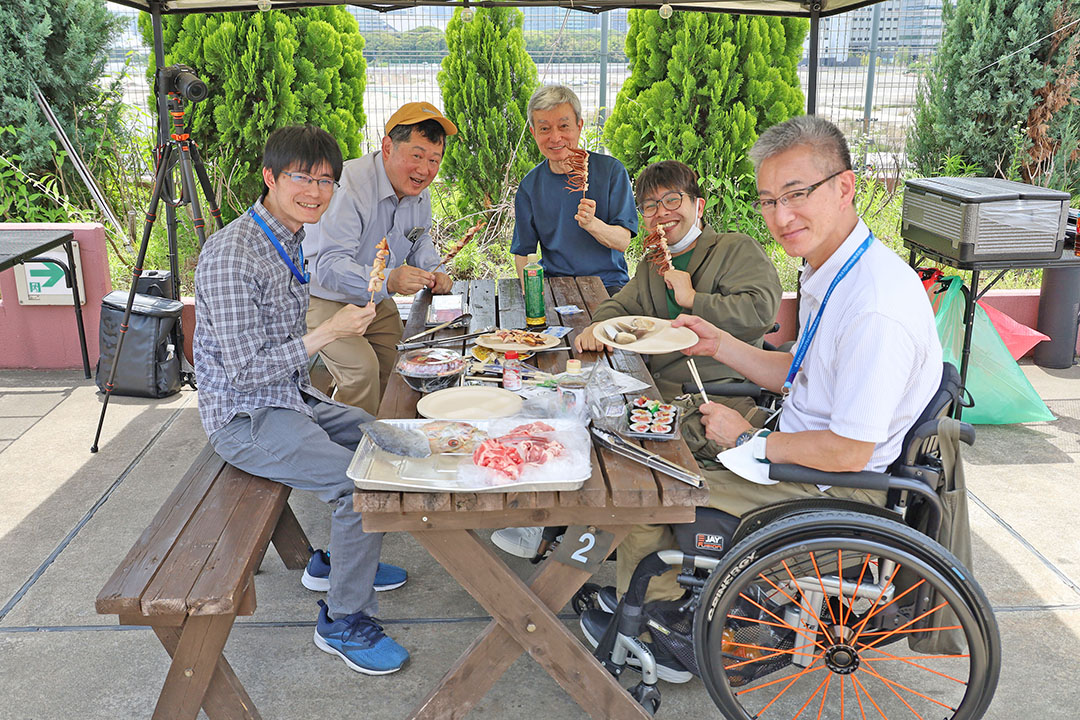 屋外のテントの下で、バーベキューやお寿司などの食事を囲みながら、談笑しリラックスした時間を過ごして楽しげです。