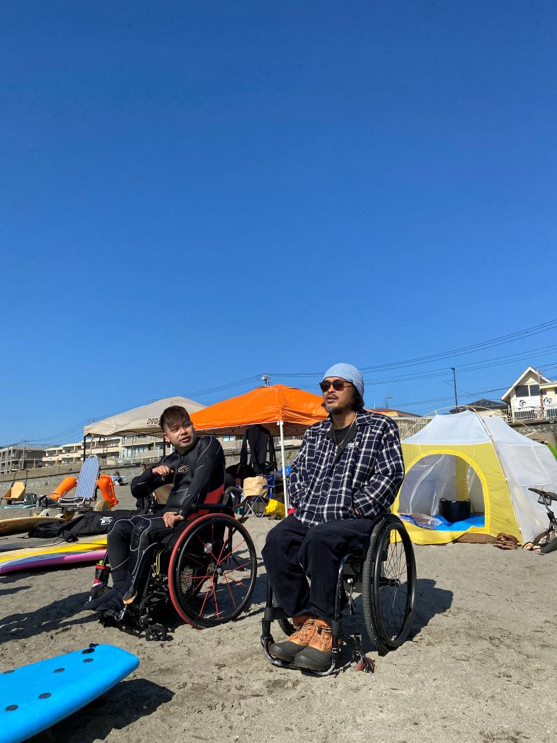 青空の砂浜で日光浴する車いすの翔太とJAHLI 後ろには小さなシェードがいくつか張られていて 多くの人がサーフィンの練習中だと思われる