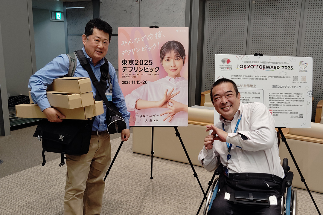 東京2025デフリンピックの告知ポスターの前で記念撮影。片方の人は荷物を持っており、もう一人は手話でメッセージを表現しています。チームワークや多様性を大切にする温かい空気が感じられます