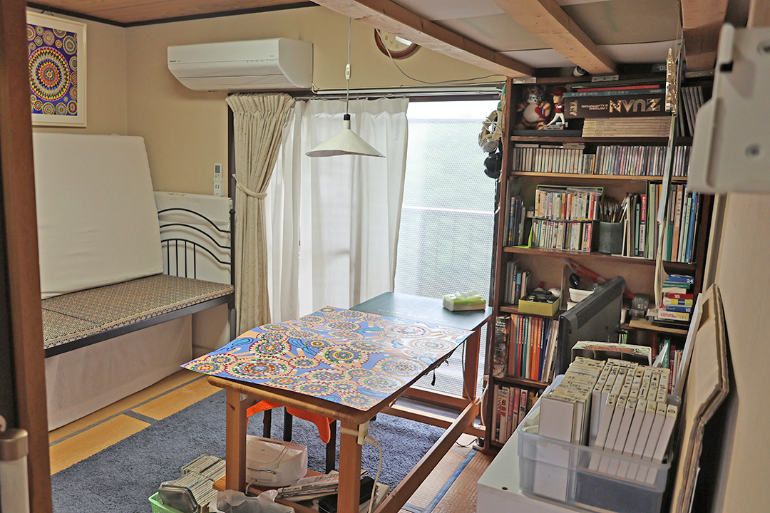 作業部屋の真ん中には1畳ほどの大きさのテーブルがあり、絵が置かれている。テレビやエアコン、本棚、ソファーがあり、快適そうな空間。