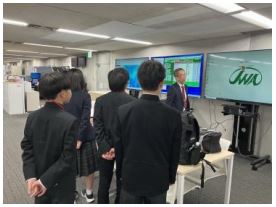 高校生くらいの男子３人、女子1人でモニター３台の前で説明を受けている様子