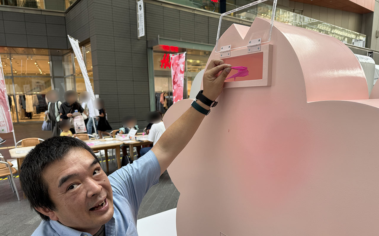 折った折り鶴をTOKYO2025モニュメントの後ろにある桜に入れているらくゆく編集者