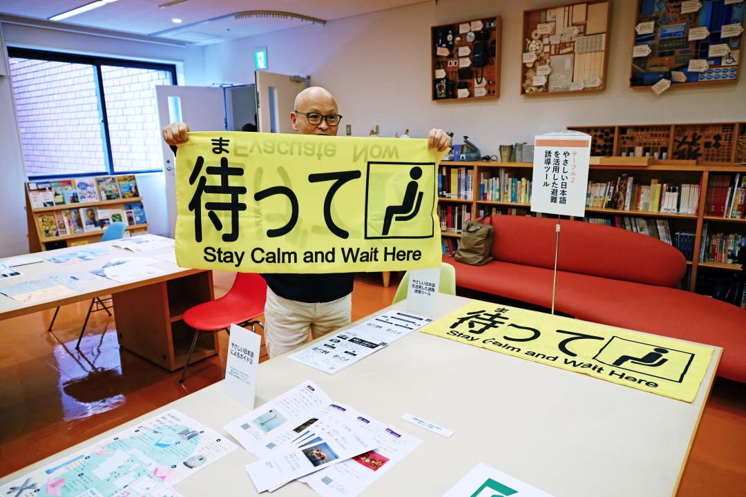 大きな黄色い旗を両手を広げてかかげる担当者「待って」と書かれている。災害時にパニックにならないようにするもの