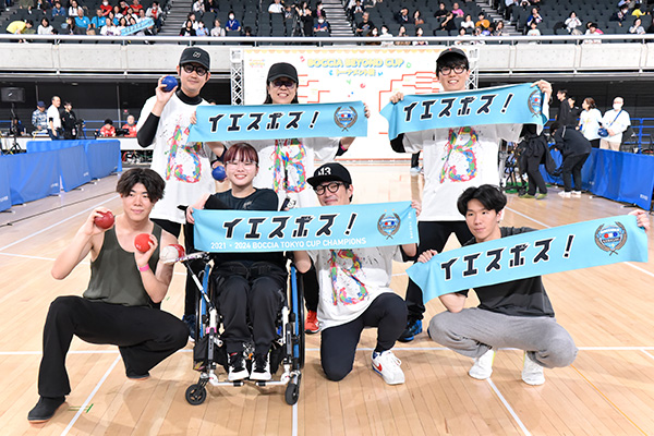 参加したチームの記念撮影。メンバーは車いすの方も含めてバナーを持ち、お揃いのTシャツを着てボッチャボールを手にしています。チームの一体感が伝わる写真です
