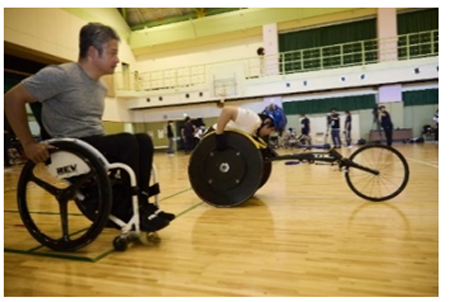 体育館でパラアスリートと陸上競技用の車いすで走っている様子。楽しそうに走っている