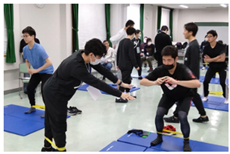 室内で複数の人がストレッチや体力測定のような運動をグループで行っている様子