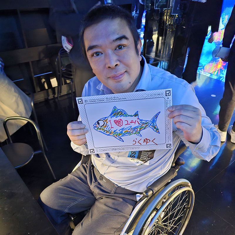 車いすの男性が描いた魚の絵を胸の前で掲げているようす