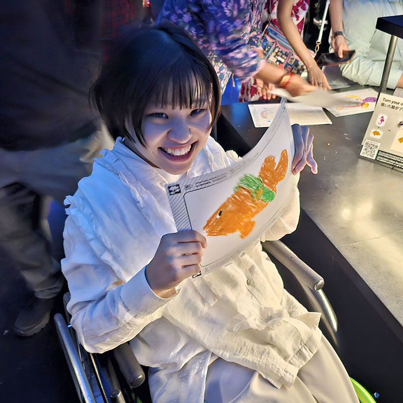 車いすの女性が描いた魚の絵を胸の前で掲げているようす