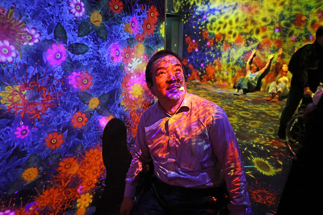 花が咲き乱れる映像の壁の前で笑顔の車いすの男性