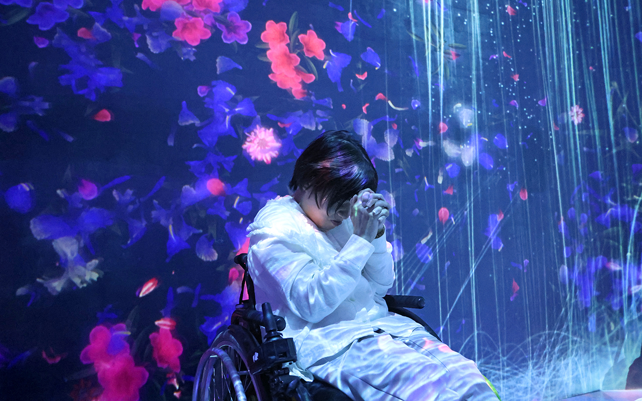 滝のような映像の壁の下で目をつぶり手を合わせて何かを祈る車いすの女性