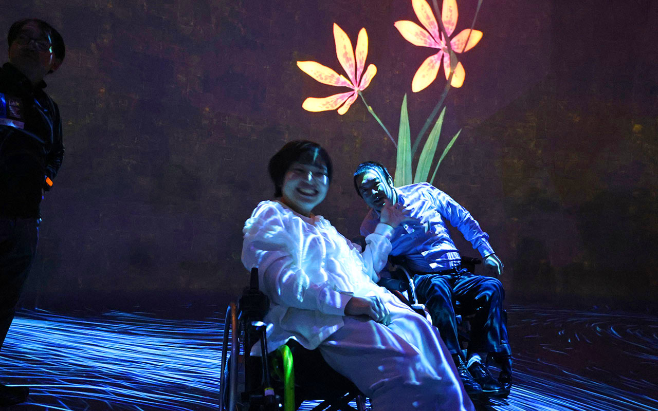 2輪咲くオレンジ色の花の映像の前で笑顔の車いすの男性と女性