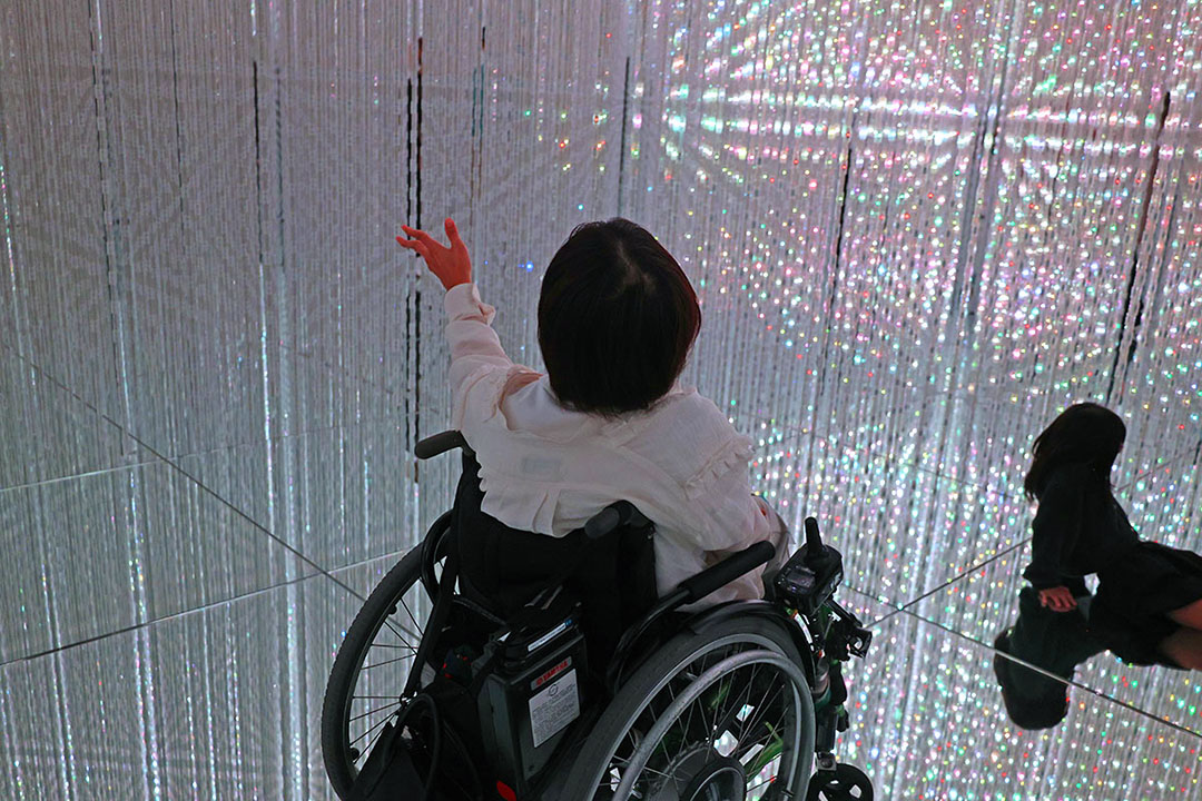 無数のクリスタル系の光の点によって構成された映像の前にいる車いすの女性