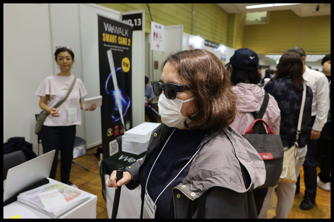 イベント会場で、遮光眼鏡とマスクを着用し、白い杖を持つ女性ライター