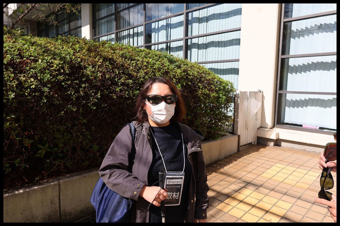 会場付近の晴れた中庭で、マスクと遮光眼鏡を着用したライターが、白い杖を持って立っている