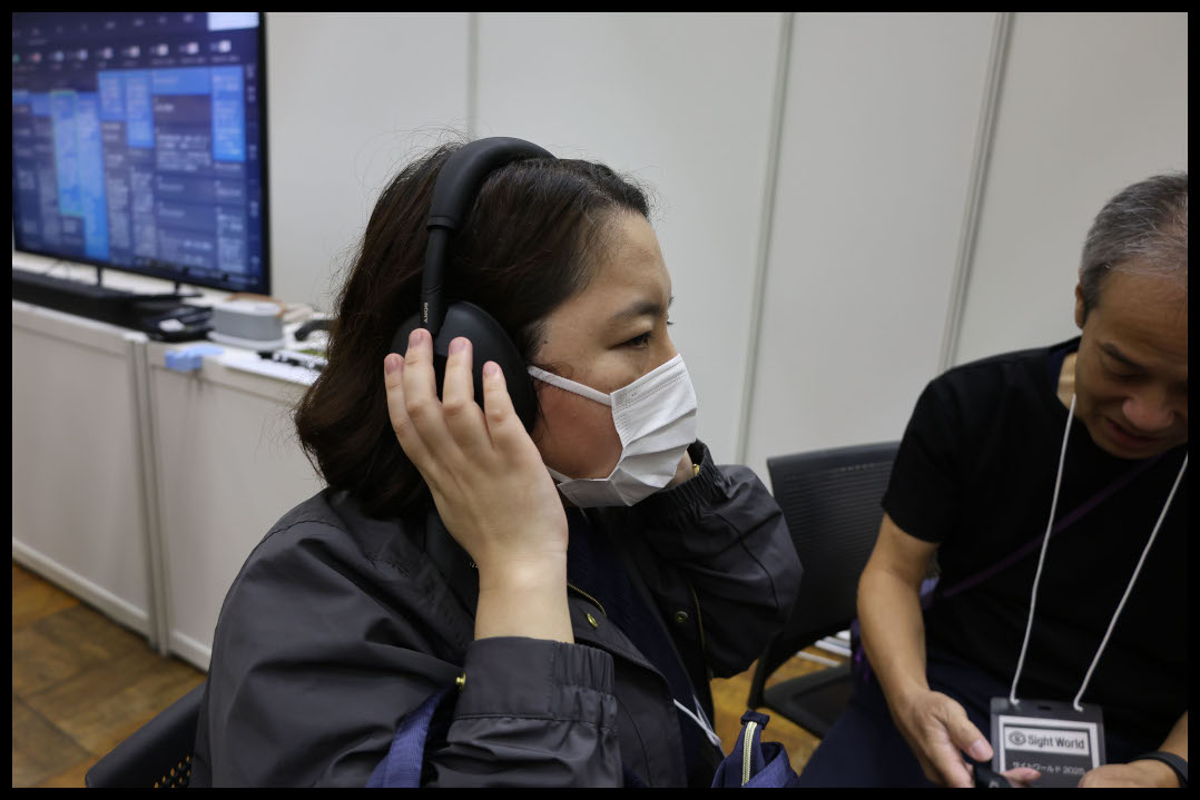 イベント会場で、ライターが黒いオーバーイヤー型ヘッドホンを試着し、音を確かめている