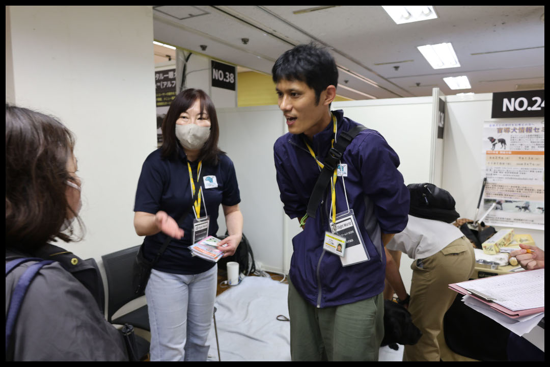 展示ブースで、黒のユニフォームを着た男性と女性のスタッフが取材に応じている様子