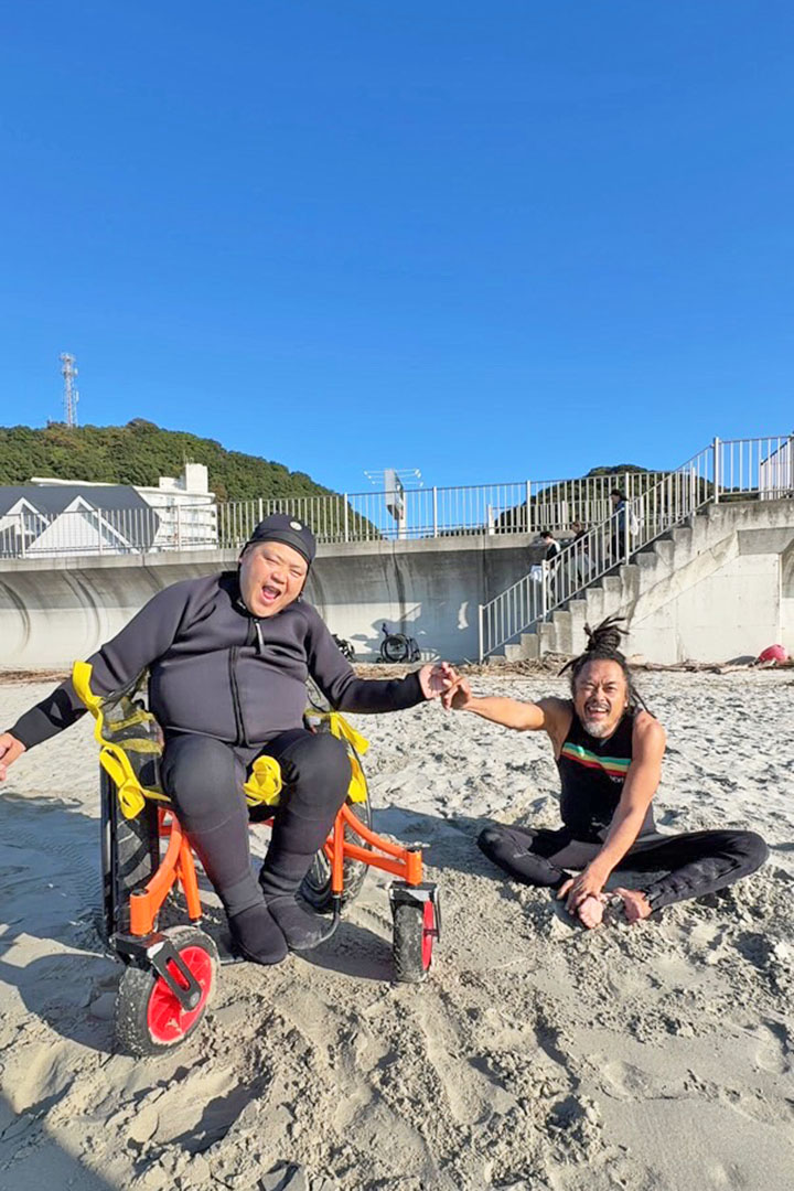 青い空の下、ビーチ用車いすに乗るダイゴロウと、砂浜にあぐらで座るジャリ