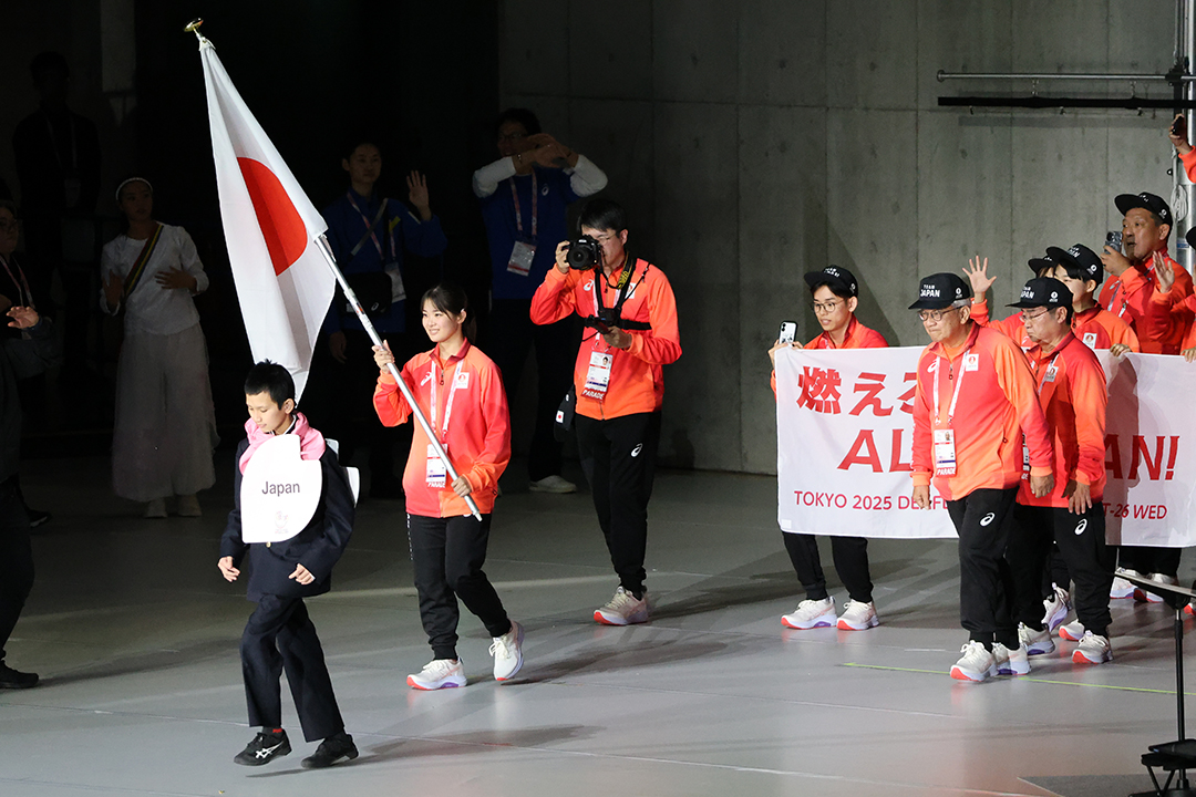 日本選手団、デフ空手の小倉 涼選手が先頭で国旗を持っています。衣装は赤系のジャージに黒いズボンです