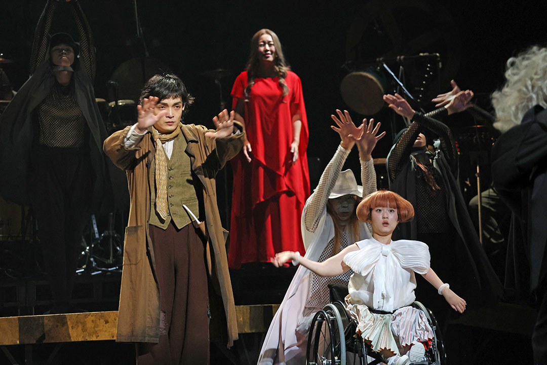 主演の男は手を前に出しています。右手前には白い衣装の車いすに乗った女性がいます