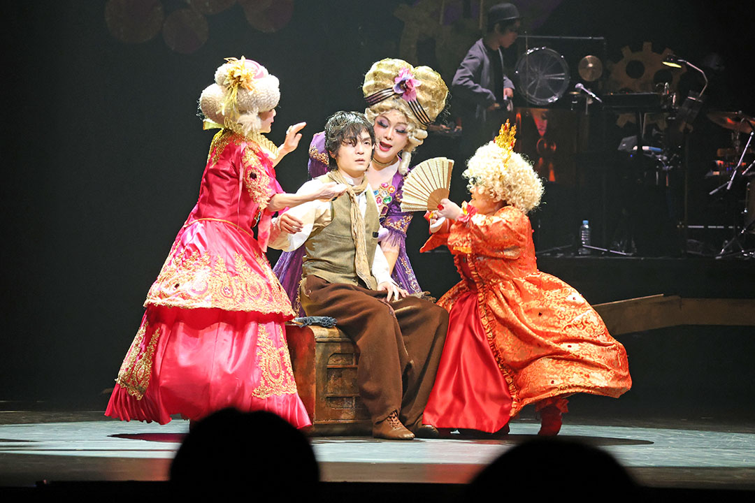 主演の男がきらびやかな衣装の３人の女性に囲まれています