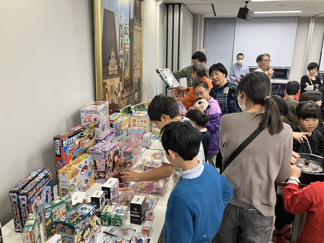 テーブルの上に並んだおもちゃに興味津々な子どもたちと、その後ろでブール盤で遊ぶ子どもたち