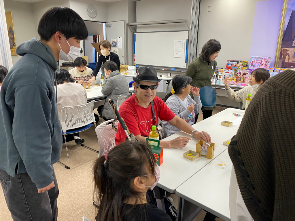 黒いハットに赤いティーシャツ、スポーティーなサングラスの出で立ちの男性の視覚障害者が笑顔で山札を引いている様子