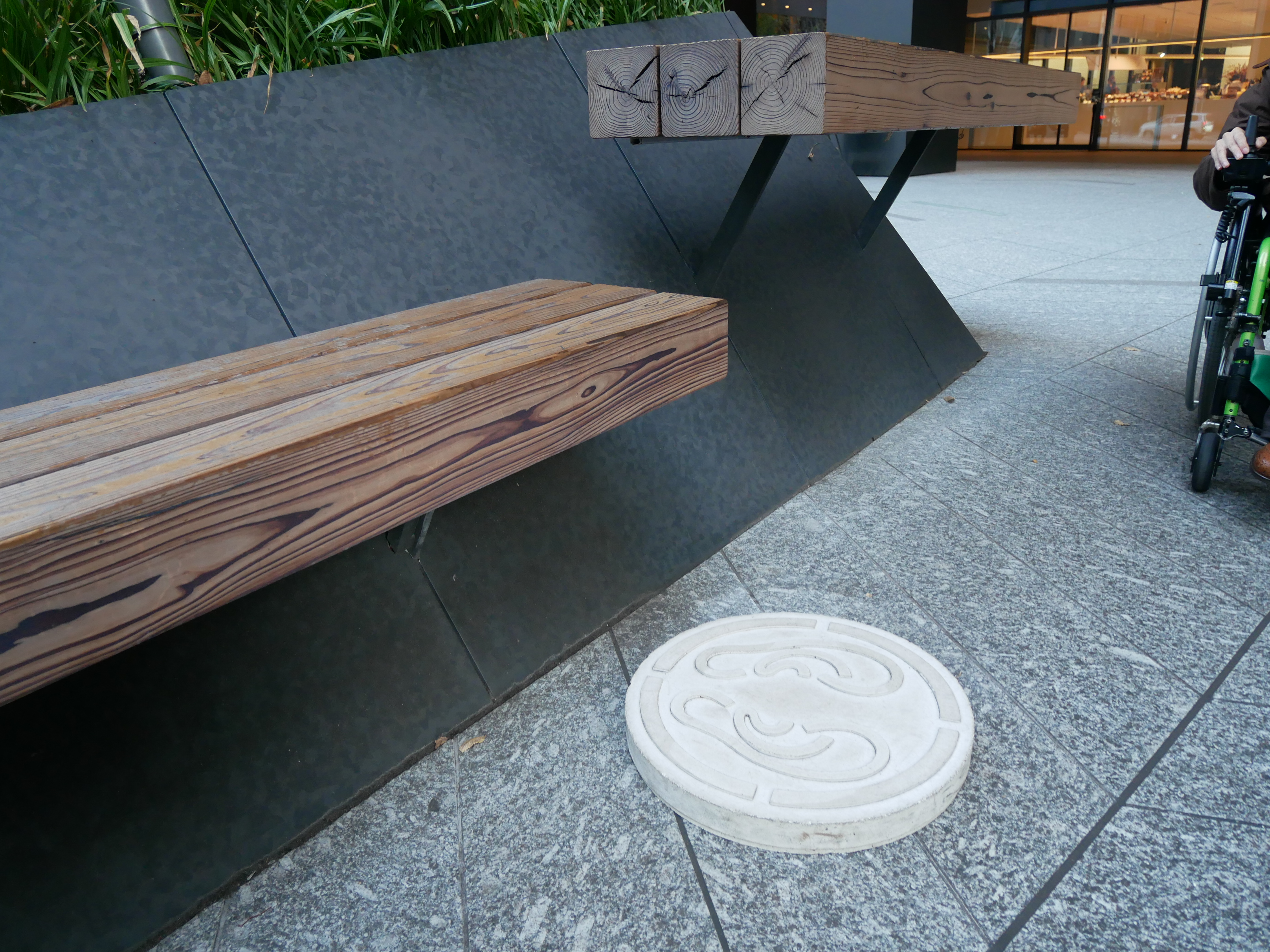 外に木製のベンチ。その足元に耳マークのようなものが描かれた白いマンホールのようなものがある。写真の右上にわずかに電動車椅子ユーザが写っている。