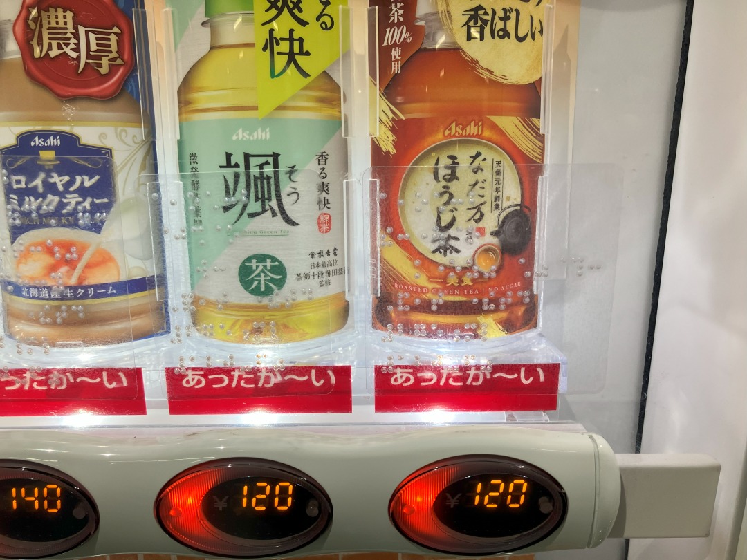 自販機をアップで写した写真。一つ一つの商品の上に透明な点字シールで商品名が書かれている